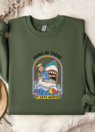 a green sweatshirt with a graphic design depicting a person in a blue shirt and a shark, along with the text "HANG IN THERE IT GETS WORSE". The sweatshirt is laid out on a bed with a white blanket and a white mug.