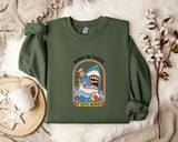 a green sweatshirt with a graphic design depicting a person in a blue shirt and a shark, along with the text "HANG IN THERE IT GETS WORSE". The sweatshirt is laid out on a bed with a white blanket and a white mug.
