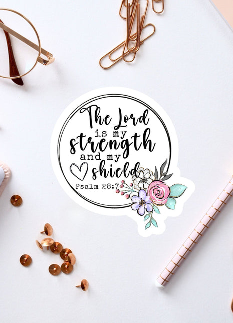 a sticker with a quote from the Bible verse Psalm 28:7, surrounded by various office supplies such as pens, a notebook, and a pair of glasses.