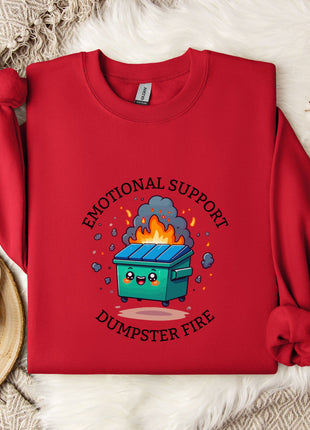 a red sweater with a graphic design depicting a dumpster with flames coming out of it, accompanied by the text "Emotional Support Dumpster Fire".