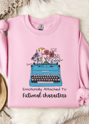 A pink sweatshirt with a graphic of a typewriter and flowers, along with the text "Emotionally Attached To Fictional Characters".