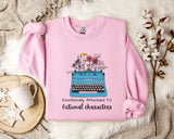 A pink sweatshirt with a graphic of a typewriter and flowers, along with the text "Emotionally Attached To Fictional Characters".