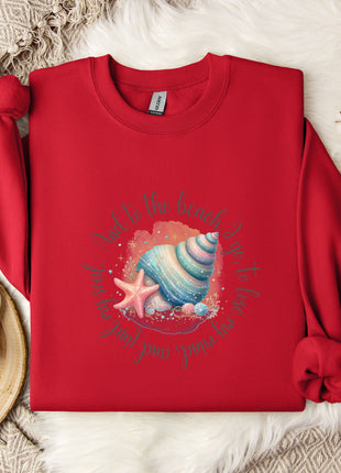 A red sweater with a colorful seashell and starfish design, along with a mug and a white flower, is displayed on a white fur rug.
