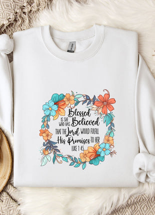 A white sweatshirt with a floral design and text printed on it, placed on a bed with a white blanket and a white mug.