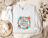 A white sweatshirt with a floral design and text printed on it, placed on a bed with a white blanket and a white mug.