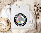 a white sweatshirt with a graphic design on it, placed on a bed with a white blanket and a mug.