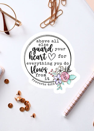 a sticker with a quote from Proverbs 4:23, surrounded by various office supplies such as pens, a notebook, and a pair of glasses.