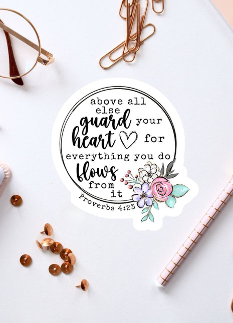 a sticker with a quote from Proverbs 4:23, surrounded by various office supplies such as pens, a notebook, and a pair of glasses.