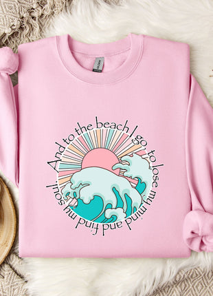 A pink sweatshirt with a graphic design of a wave and sun, along with the text "And to the beach I go to lose my mind" printed on it, is displayed on a bed with a white blanket and a mug.