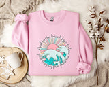 A pink sweatshirt with a graphic design of a wave and sun, along with the text "And to the beach I go to lose my mind" printed on it, is displayed on a bed with a white blanket and a mug.