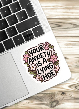 A sticker with the text "Your anxiety is a lying hoe" is placed on the edge of a laptop keyboard.