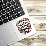 A sticker with the text "Your anxiety is a lying hoe" is placed on the edge of a laptop keyboard.