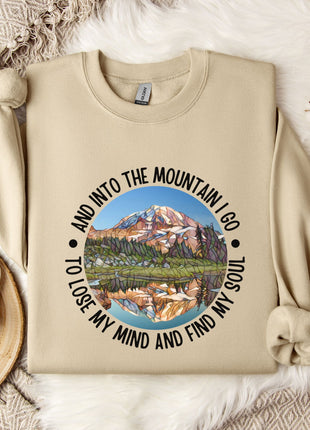 A beige sweatshirt with a mountain landscape design and the text "And into the mountain I go to lose my mind and find my soul" printed on it, along with a mug and some flowers on a bed.