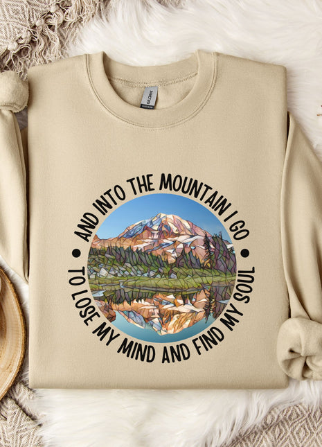 A beige sweatshirt with a mountain landscape design and the text "And into the mountain I go to lose my mind and find my soul" printed on it, along with a mug and some flowers on a bed.