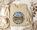 A beige sweatshirt with a mountain landscape design and the text "And into the mountain I go to lose my mind and find my soul" printed on it, along with a mug and some flowers on a bed.