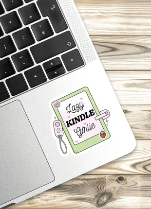 A sticker with a cartoon-style illustration of a book and the text "Lazy Kindle Girlie" is placed on the edge of a laptop keyboard.