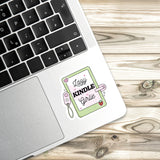 A sticker with a cartoon-style illustration of a book and the text "Lazy Kindle Girlie" is placed on the edge of a laptop keyboard.
