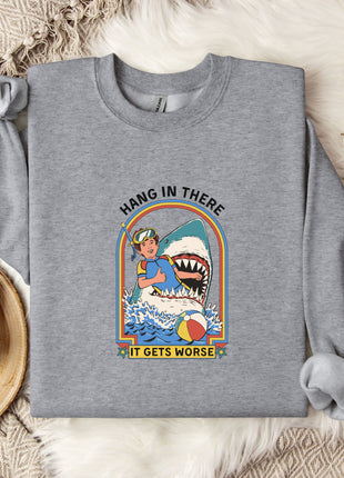 A gray sweatshirt with a graphic design depicting a person in a blue outfit and a shark, along with the text "HANG IN THERE IT GETS WORSE". The sweatshirt is laid out on a bed with a white blanket and a mug.