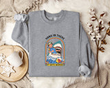 A gray sweatshirt with a graphic design depicting a person in a blue outfit and a shark, along with the text "HANG IN THERE IT GETS WORSE". The sweatshirt is laid out on a bed with a white blanket and a mug.