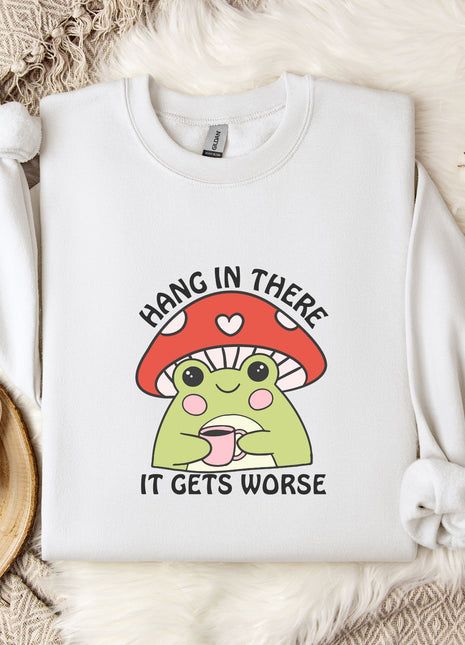 A white sweatshirt with a mushroom graphic and the text "HANG IN THERE IT GETS WORSE" printed on it, along with a mug and a branch of cotton bolls.