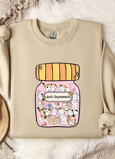 a beige sweater with a graphic design of a jar filled with various cartoon animals, including cats and dogs, and the text "Anti-Depressants" printed on it. The sweater is laid out on a bed with a white blanket and a small white mug, along with some white flowers.