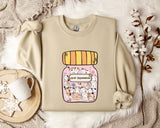 a beige sweater with a graphic design of a jar filled with various cartoon animals, including cats and dogs, and the text "Anti-Depressants" printed on it. The sweater is laid out on a bed with a white blanket and a small white mug, along with some white flowers.
