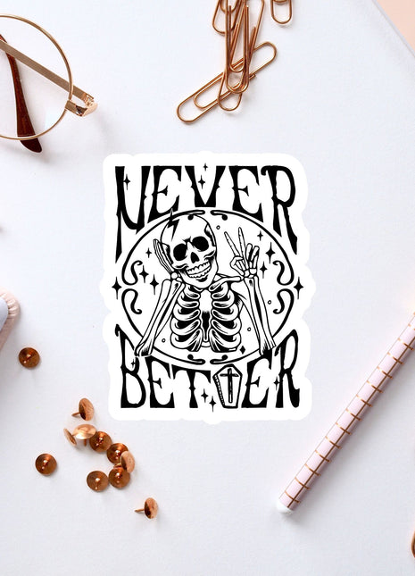 a sticker with a skeleton and the text "NEVER BETTER" on a white background, surrounded by various office supplies such as a pen, a pair of glasses, and a notebook.