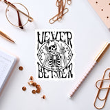 a sticker with a skeleton and the text "NEVER BETTER" on a white background, surrounded by various office supplies such as a pen, a pair of glasses, and a notebook.