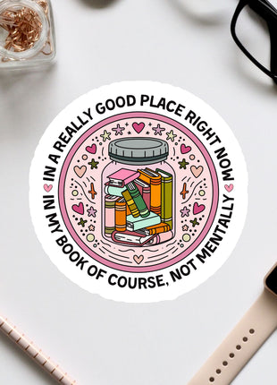 A sticker with a book inside a jar surrounded by various decorative elements, along with a pair of glasses, a pen, and a watch on a white surface.