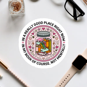 A sticker with a book inside a jar surrounded by various decorative elements, along with a pair of glasses, a pen, and a watch on a white surface.