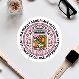 A sticker with a book inside a jar surrounded by various decorative elements, along with a pair of glasses, a pen, and a watch on a white surface.